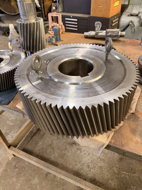 Large helical gear with lifting eyes; a pinion shaft is visible in the background.